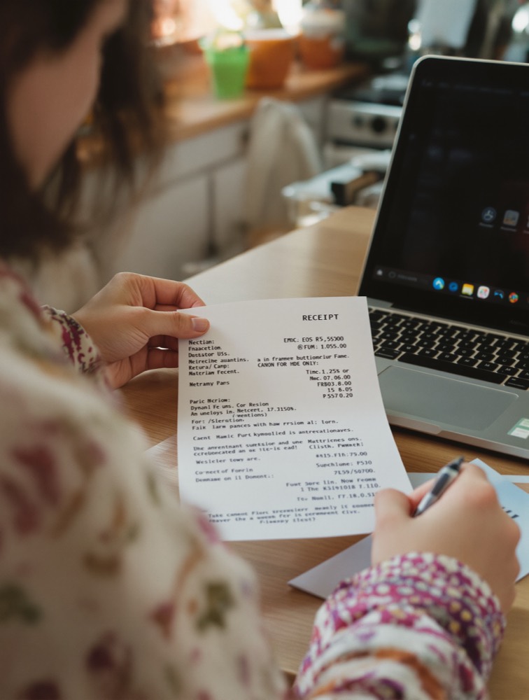 A woman at a kitchen table reading a printed receipt, her laptop and a notepad beside her.
